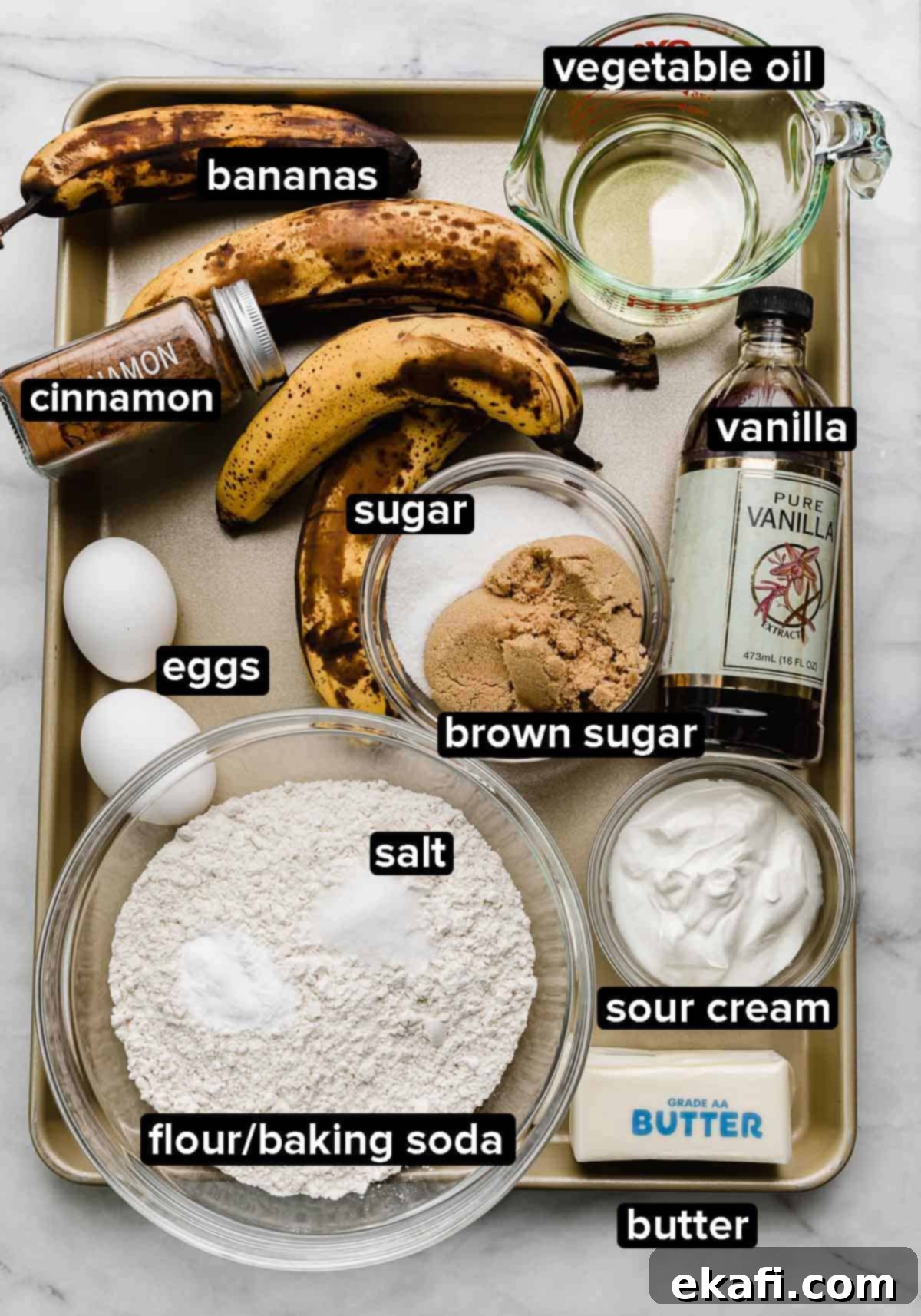 Classic Banana Bread Bundt Cake 3 Banana Bread Bundt Cake ingredients on a gold baking sheet: overly ripe bananas, sour cream, butter, eggs, oil, vanilla, sugars, and other dry ingredients.