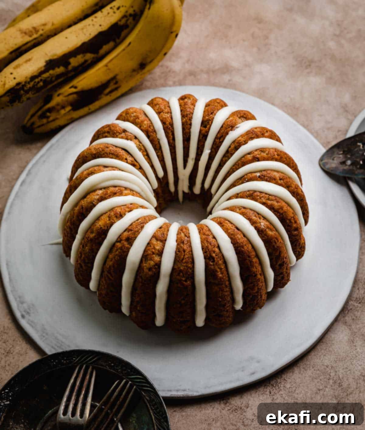Classic Banana Bread Bundt Cake 7 A cream cheese glazed banana bundt cake on a white cake plate that's on a brown textured background.