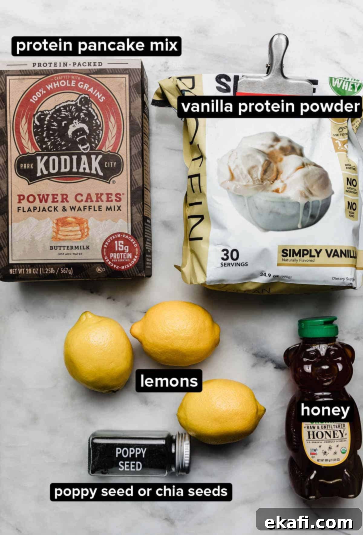 Zesty Lemon Protein Bites 3 Lemon Protein Ball ingredients on a white marble surface: vanilla protein powder, whole lemons, honey, protein pancake mix, and poppy seeds.