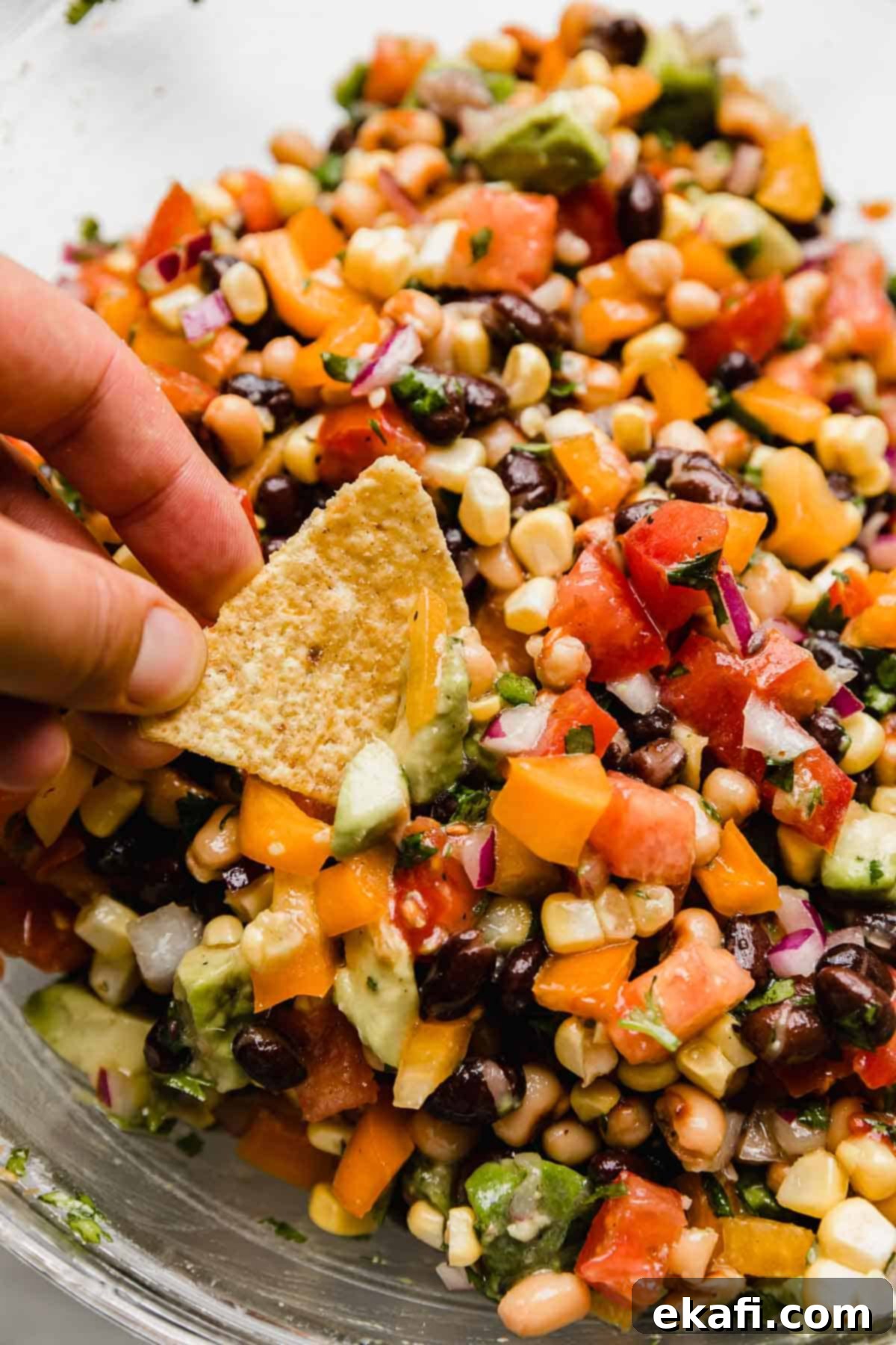 Συνταγή για Καουμπόι Χαβιάρι 2 Texas Caviar (κομμένες ντομάτες, πιπεριές, φασόλια, κόλιανδρο και κρεμμύδι) σε ένα γυάλινο μπολ με ένα χέρι να βουτά ένα τσιπ τορτίγιας στο ντιπ Cowboy Caviar.