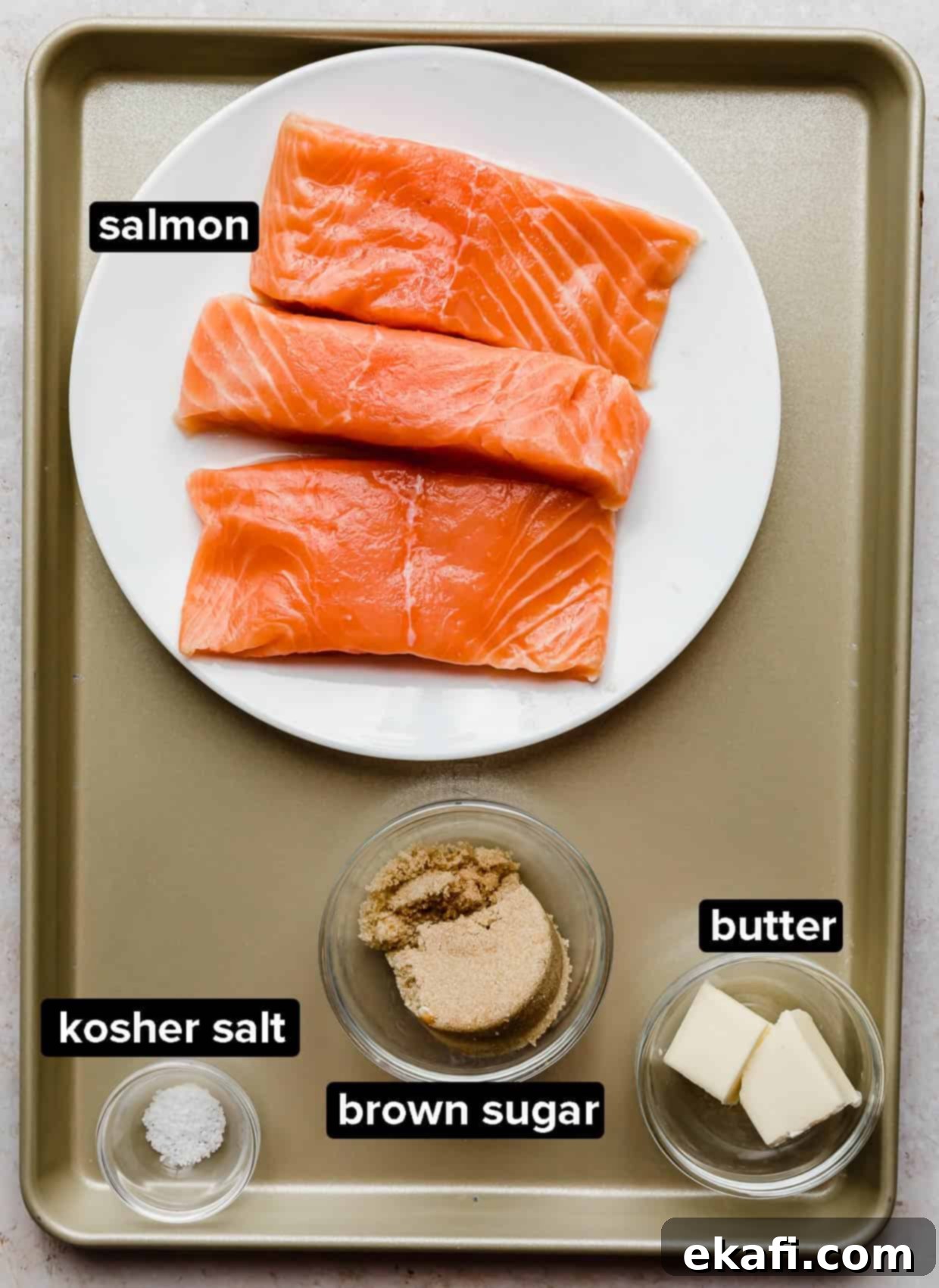 Caramelized Brown Sugar Salmon 3 A collection of fresh ingredients for Brown Sugar Salmon, including salmon fillets, butter, brown sugar, and kosher salt, arranged on a rustic bronze baking sheet.