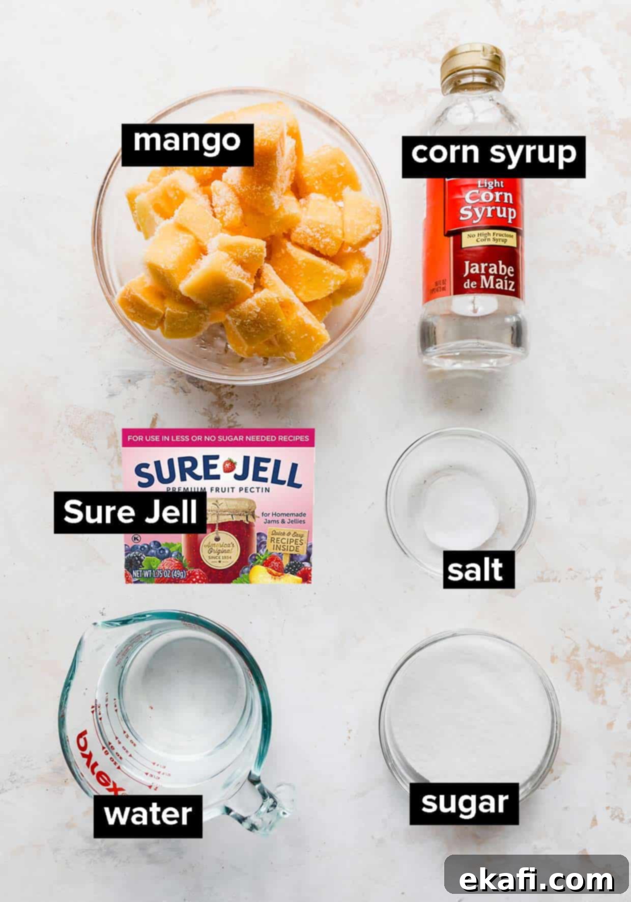 Tropical Mango Freeze 3 A flat lay image displaying the ingredients used to make mango sorbet: fresh mango chunks, light corn syrup, granulated sugar, Sure-Jell, salt, and water, all arranged on a white textured background.