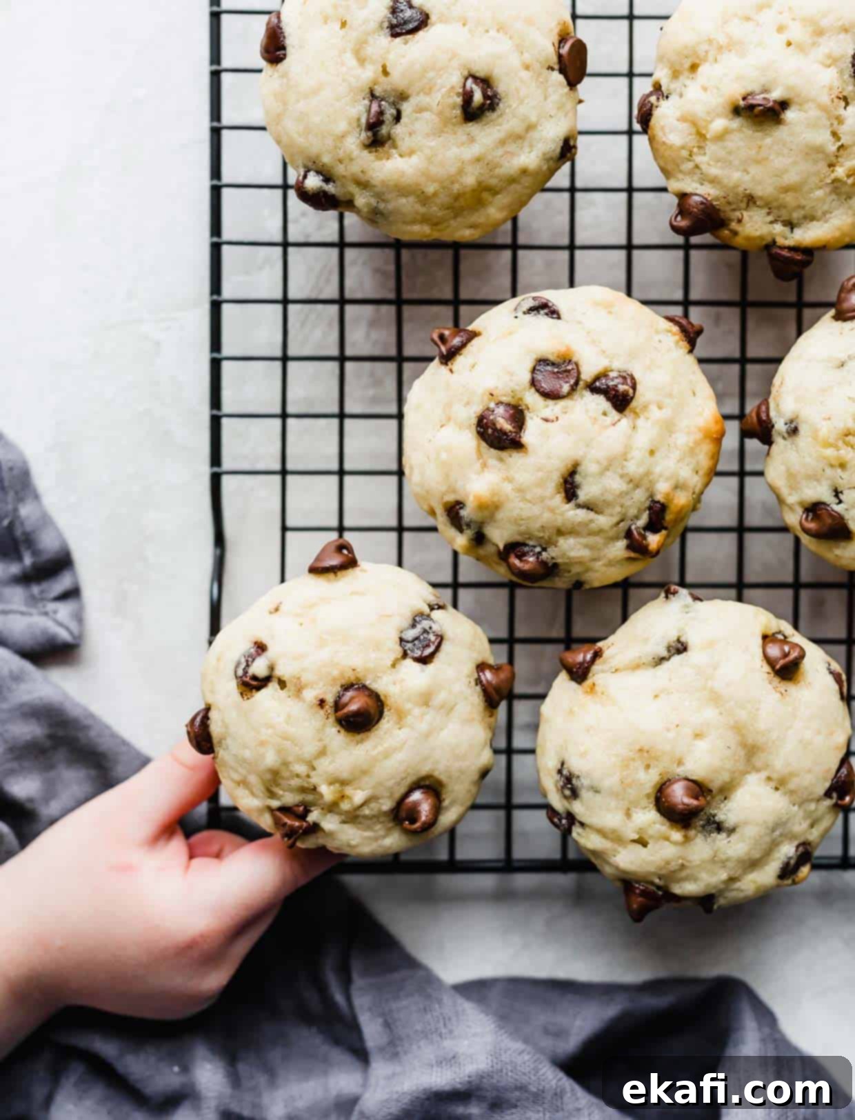 Velvet Banana Chocolate Chip Muffins 8 A small hand grabbing a chocolate chip muffin made with bananas on a wire rack.