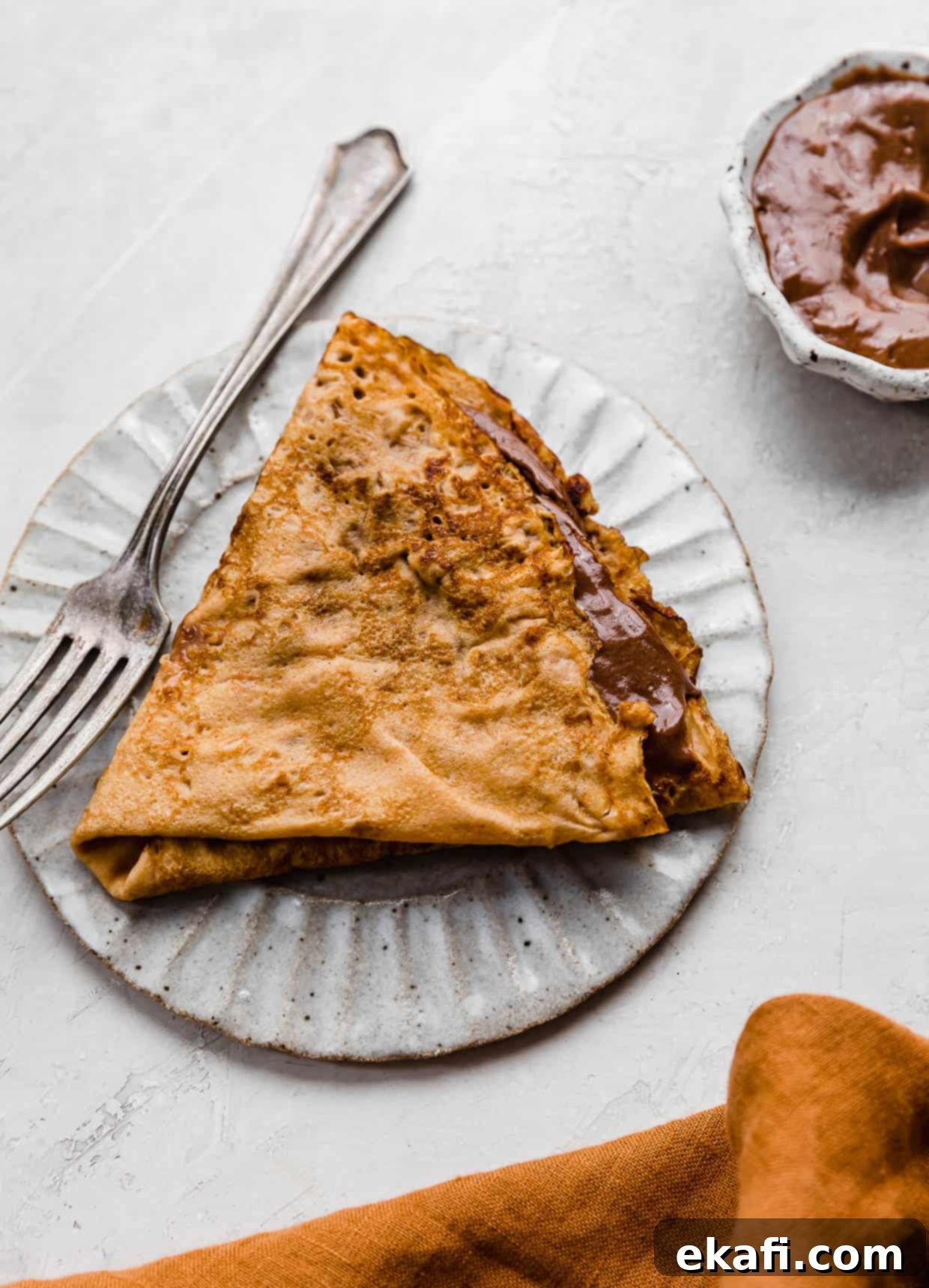 Autumn Pumpkin Crepes 2 A Pumpkin Crepe filled with whipped nutella folded into a triangle, on a white plate.