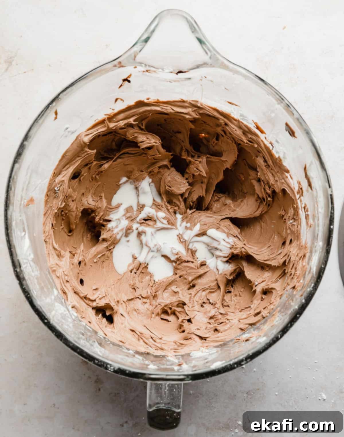 Decadent Chocolate Buttercream 12 Heavy cream being poured over light brown Chocolate Buttercream Frosting in a glass bowl, ready for final mixing.