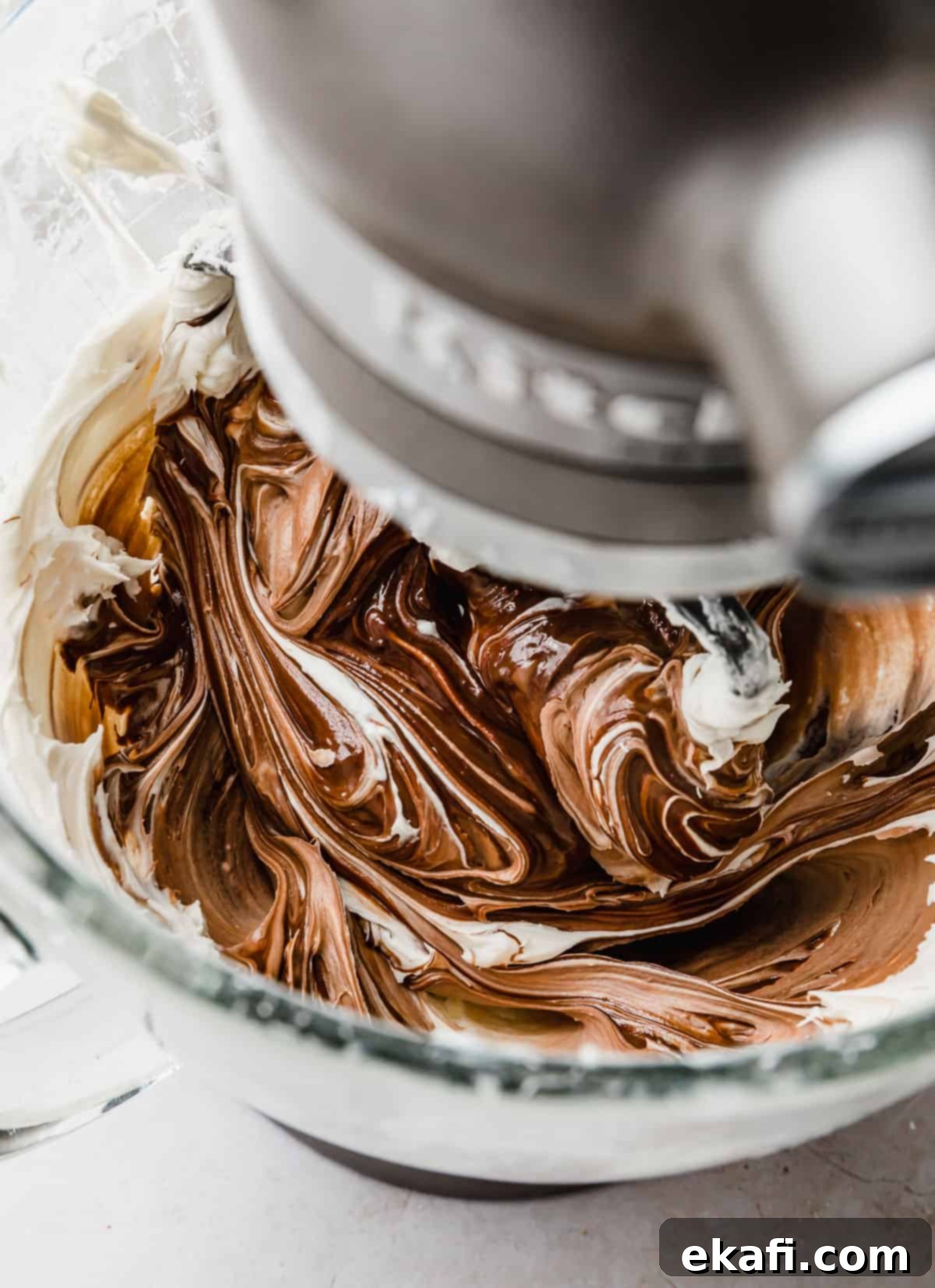 Decadent Chocolate Buttercream 9 Chocolate Buttercream Frosting with Melted Chocolate actively being mixed in a glass bowl of a stand mixer, demonstrating its evolving texture.