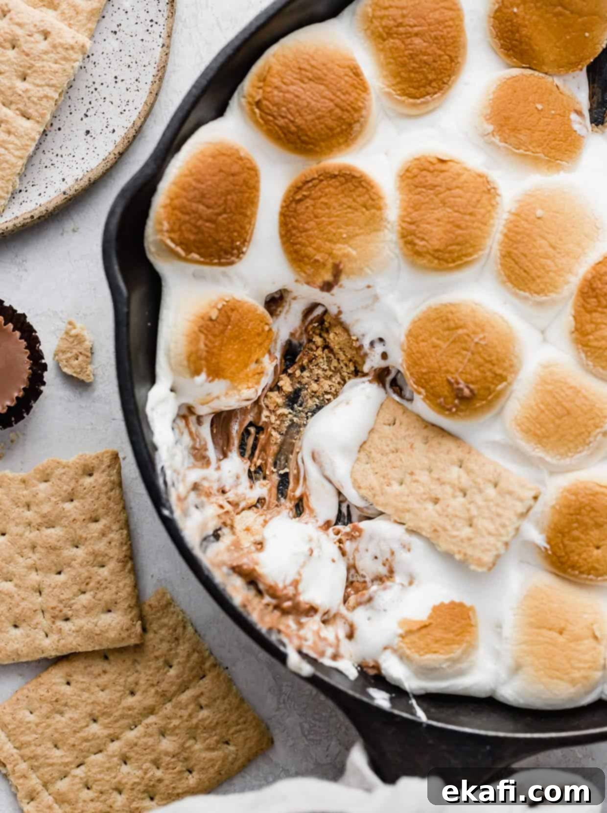 Ντιπ Σμόρες Ρήσης 2 Λαχταριστή Reese's S'mores Dip σε μαντεμένιο τηγάνι με ένα κράκερ Graham να βουτάει μέσα σε λιωμένα marshmallows και κύπελλα φυστικοβούτυρου.