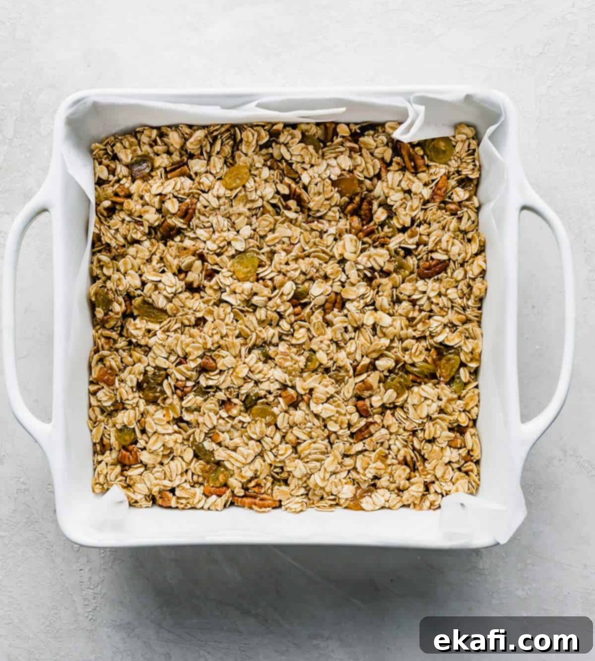 Μπάρες Γκρανόλα Βρώμης Σταφίδας 7 A white square baking dish filled with an Oatmeal Raisin Granola Bar mixture, o na white background.