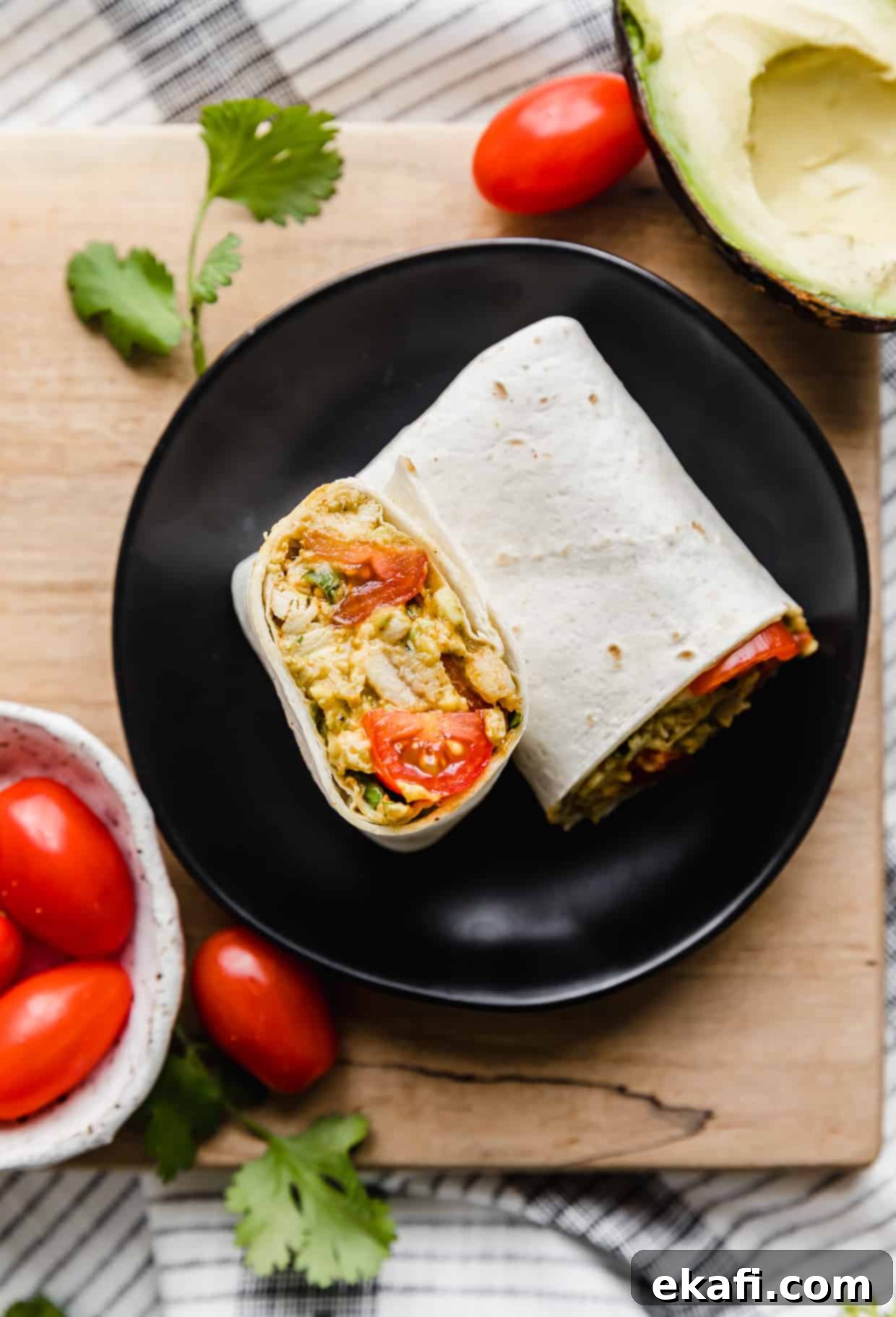Creamy Avocado Chicken Wraps 3 An Avocado Chicken Wrap on a black plate with grape tomatoes and sliced avocado surrounding it, highlighting fresh ingredients.