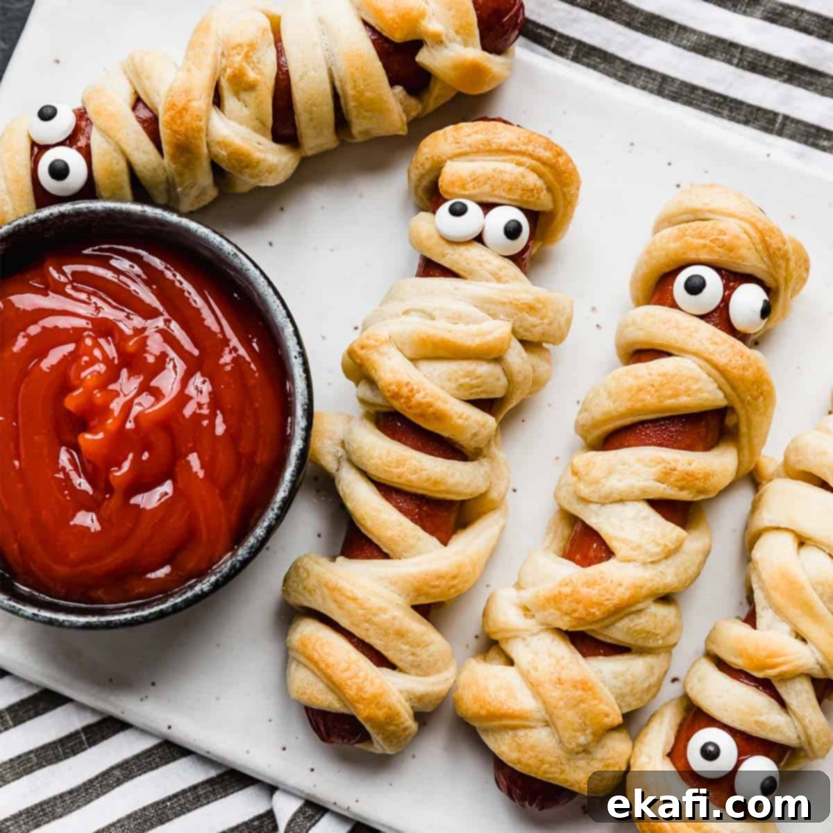 Happy Tummies Happy Kids 16 Playful Mummy Hot Dogs arranged on a white plate, with a small bowl of ketchup to the left, ready for a fun and spooky Halloween meal.