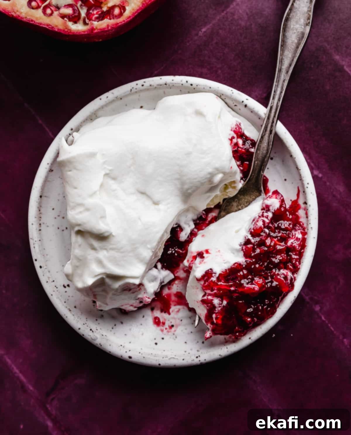 Ruby Pomegranate Delight 2 A slice of whipped cream topped Pomegranate Jell-O salad on a white plate on a deep maroon colored background.