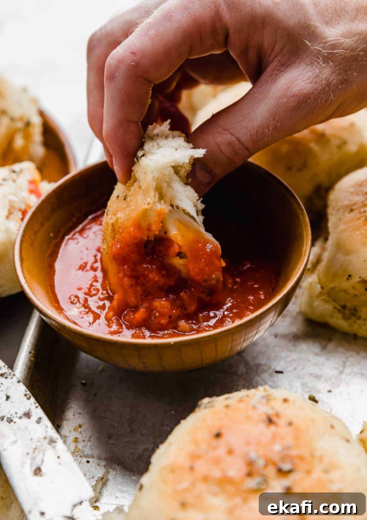 From-Scratch Pizza Rolls 7 A hand dipping a golden-brown pepperoni pizza roll into a bowl of vibrant red pizza sauce.