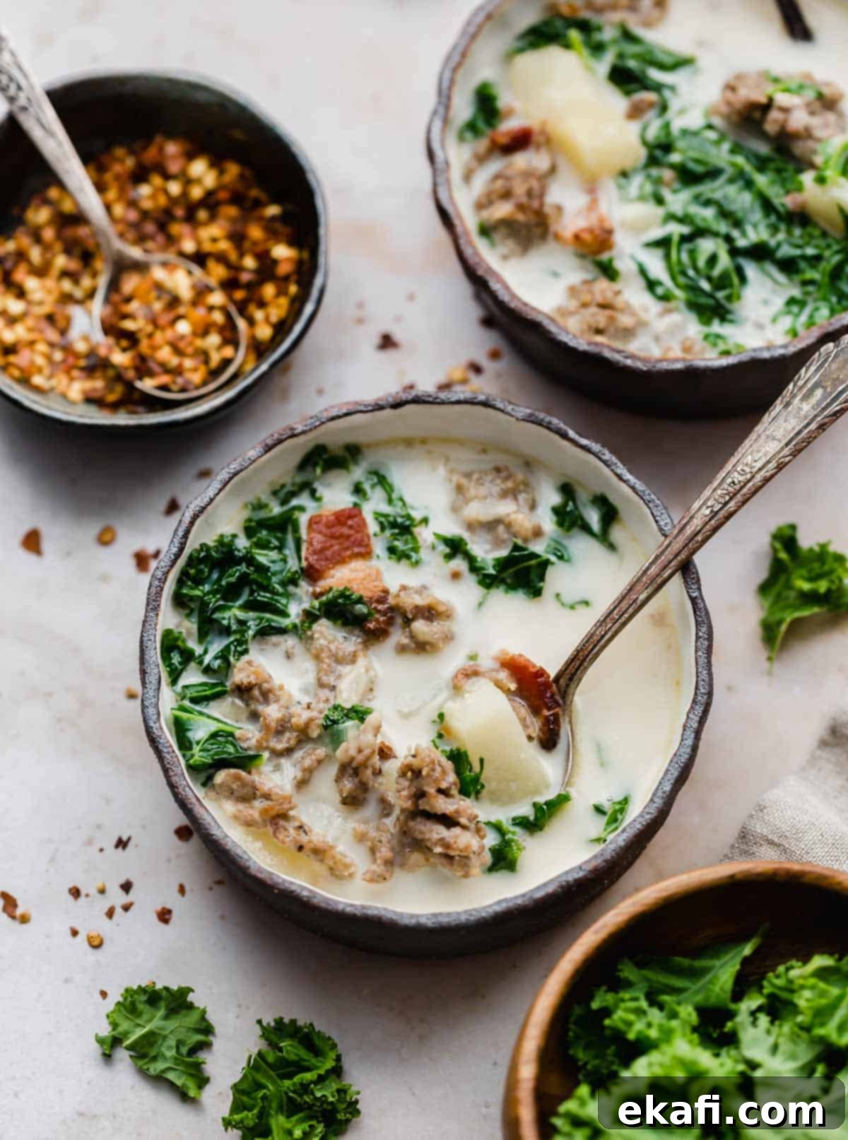 Copycat Olive Garden Zuppa Toscana 5 Olive Garden Zuppa Toscana Soup recipe in a black rimmed bowl on a cream textured background with a black bowl filled with crushed red pepper flakes in the background.