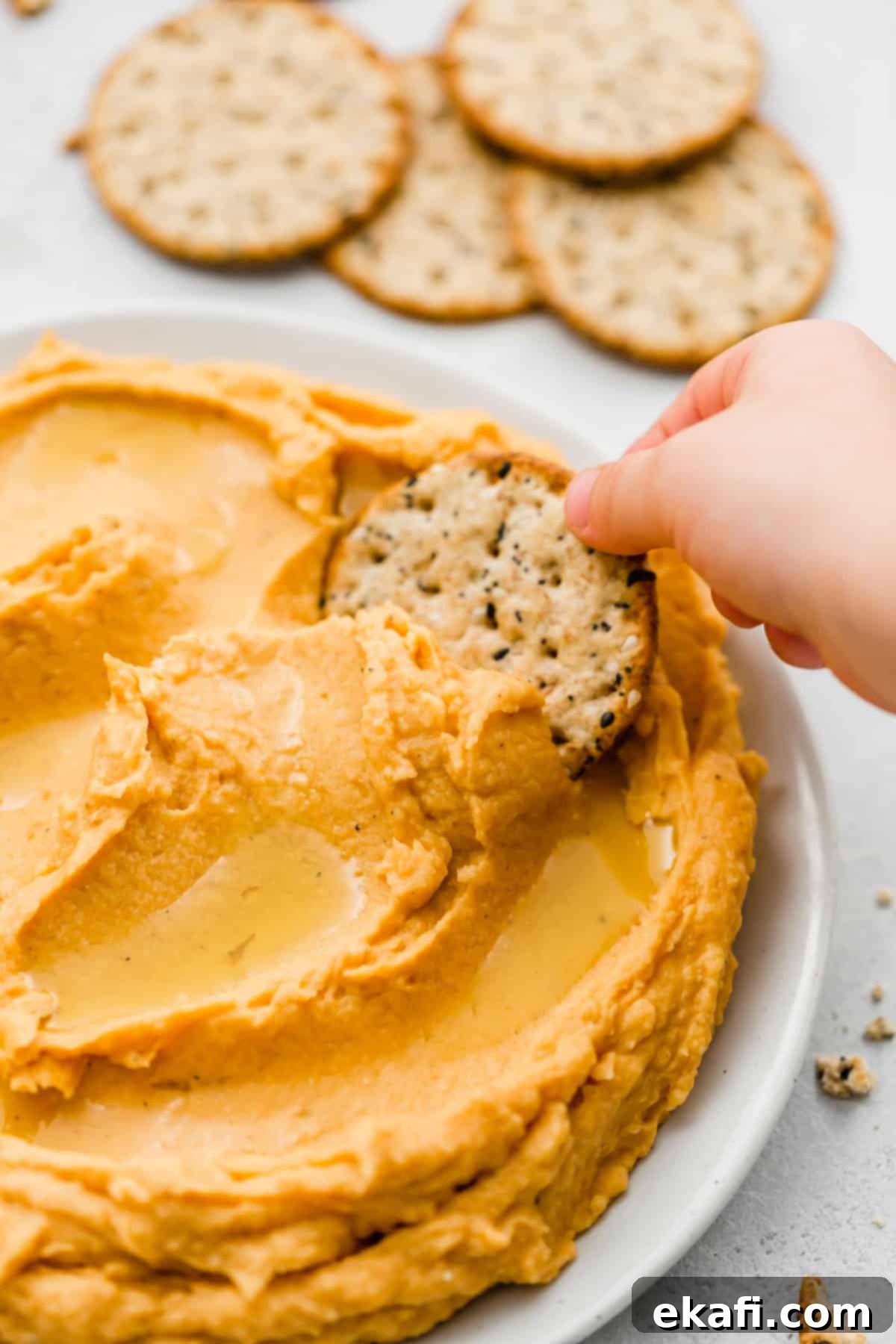 Sweet Potato Hummus 2 A hand dipping a round cracker into orange Sweet Potato Hummus, showcasing its creamy texture.