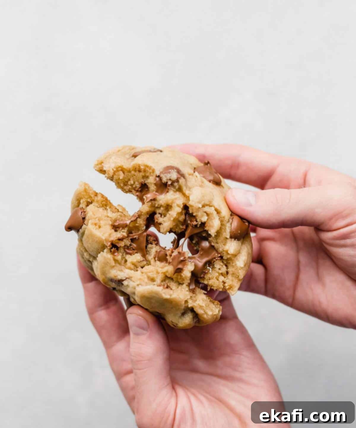 Copycat Crumbl Chocolate Chip Cookie Perfection 11 A pair of male hands holding a large, freshly baked Crumbl Chocolate Chip Cookie and splitting it in half, revealing its gooey, melty chocolate interior.