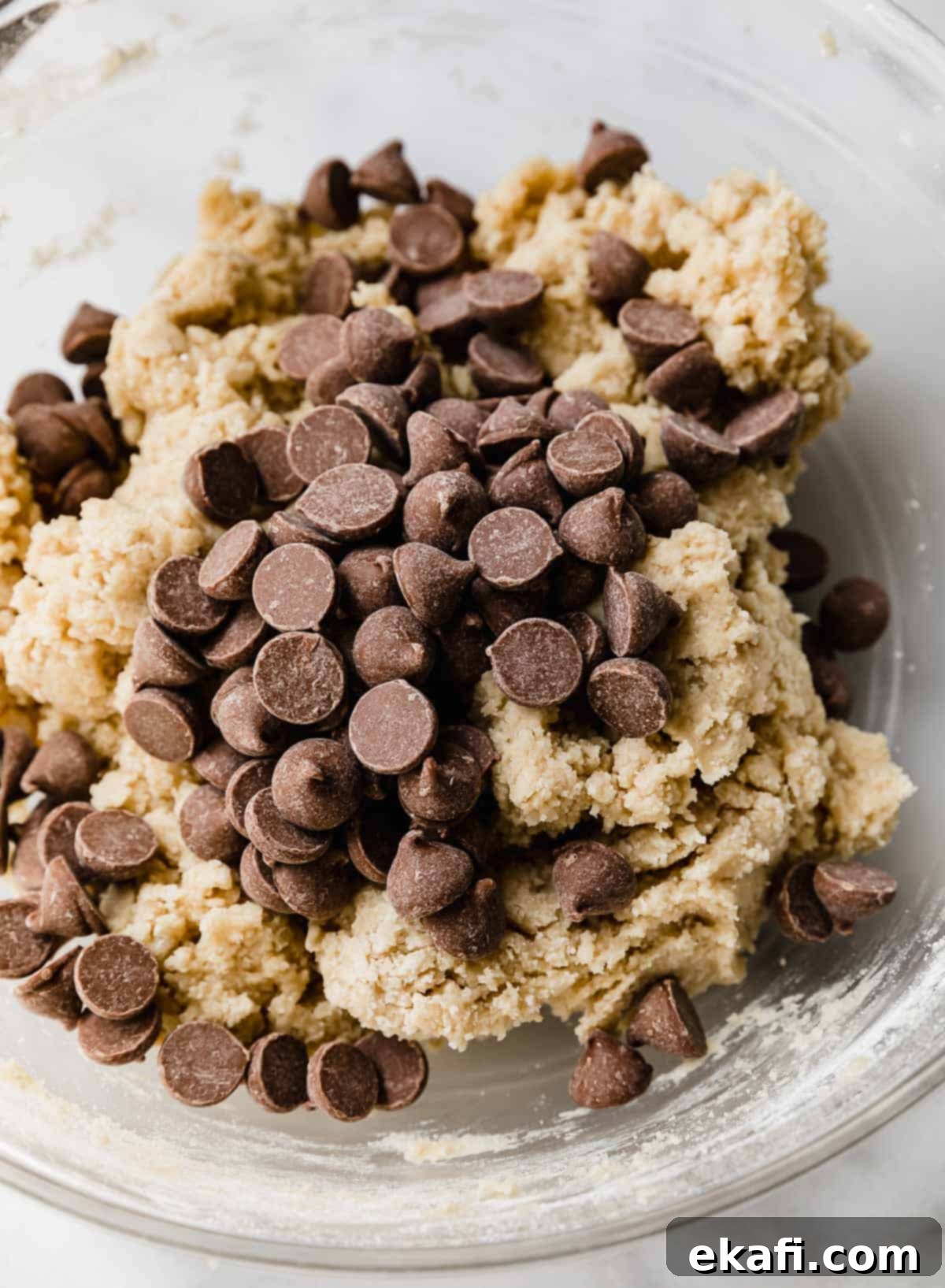 Copycat Crumbl Chocolate Chip Cookie Perfection 7 Milk chocolate chips sprinkled over homemade Crumbl chocolate chip cookie dough in a glass bowl, ready for mixing.