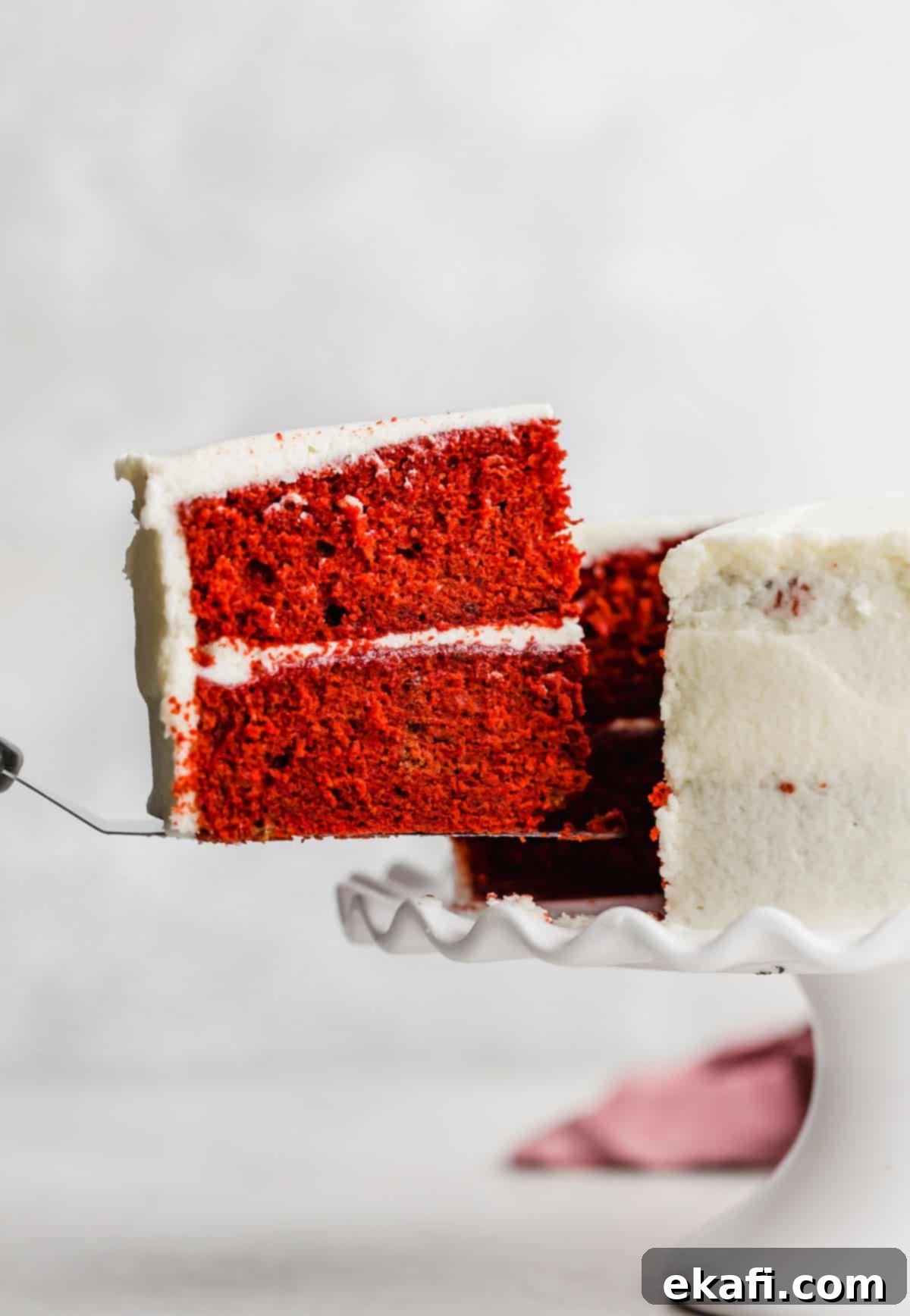Heirloom Red Velvet Cake 2 A perfectly sliced piece of Old Fashioned Red Velvet Cake being lifted from a freshly frosted cake, showcasing its vibrant red layers and delicate white icing.