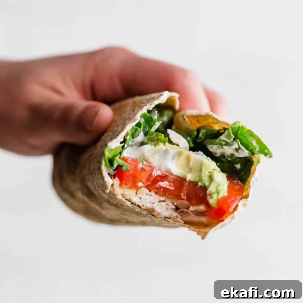 Υγιεινές Τυλιχτές Κοτόπουλο 6 Ένα χέρι κρατά μια Υγιεινή Τυλιχτή Κοτόπουλου με μια μπουκιά που έχει αφαιρεθεί από την τυλιχτή, δείχνοντας το αβοκάντο, τη ντομάτα, το κοτόπουλο και το σπανάκι στην τυλιχτή.