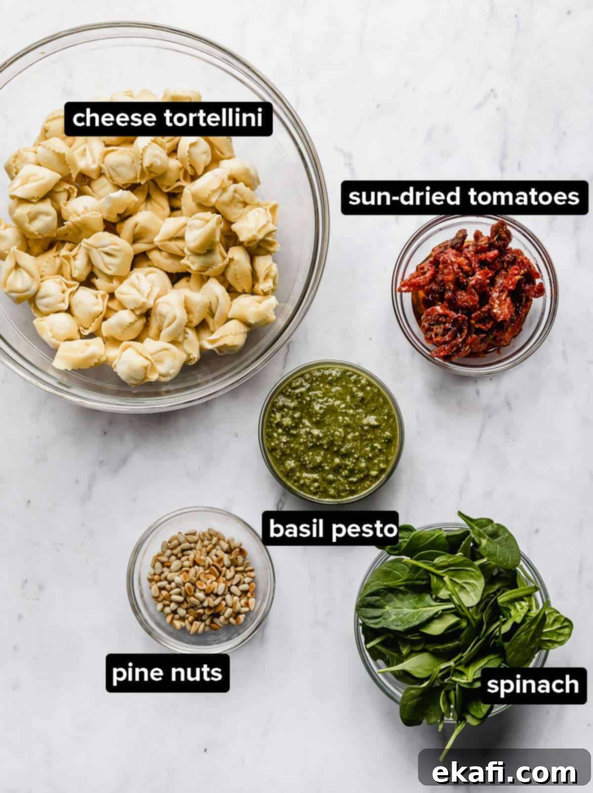 Pesto-Kissed Tortellini 3 Ingredients used to make Pesto Tortellini Pasta Salad, portioned into glass bowls: tortellini, pesto, sun-dried tomatoes, pine nuts, and optional spinach.