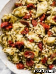 Pesto-Kissed Tortellini 6 A close up photo of Pesto Tortellini Pasta Salad tossed with sun-dried tomatoes and pine nuts in a white bowl.