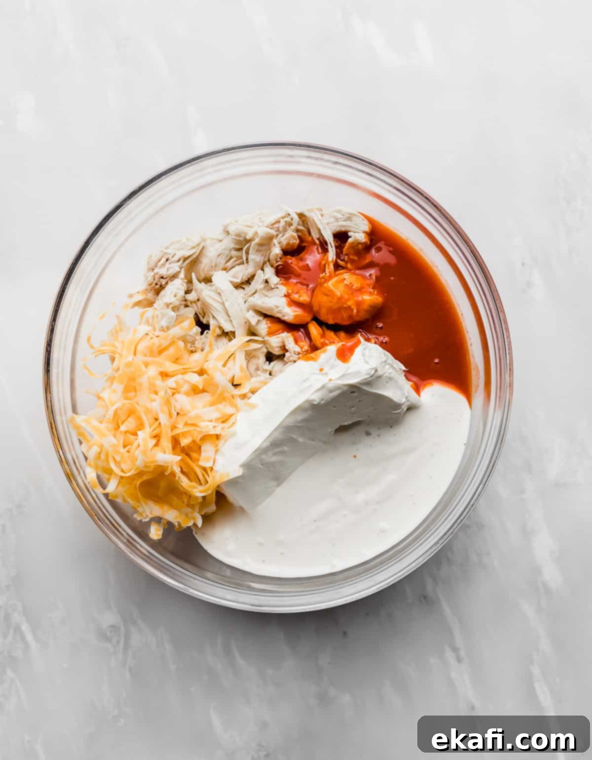 Ultimate Buffalo Chicken Dip 4 A glass bowl with shredded chicken, cream cheese, buffalo sauce, ranch dressing, and cheese in it.
