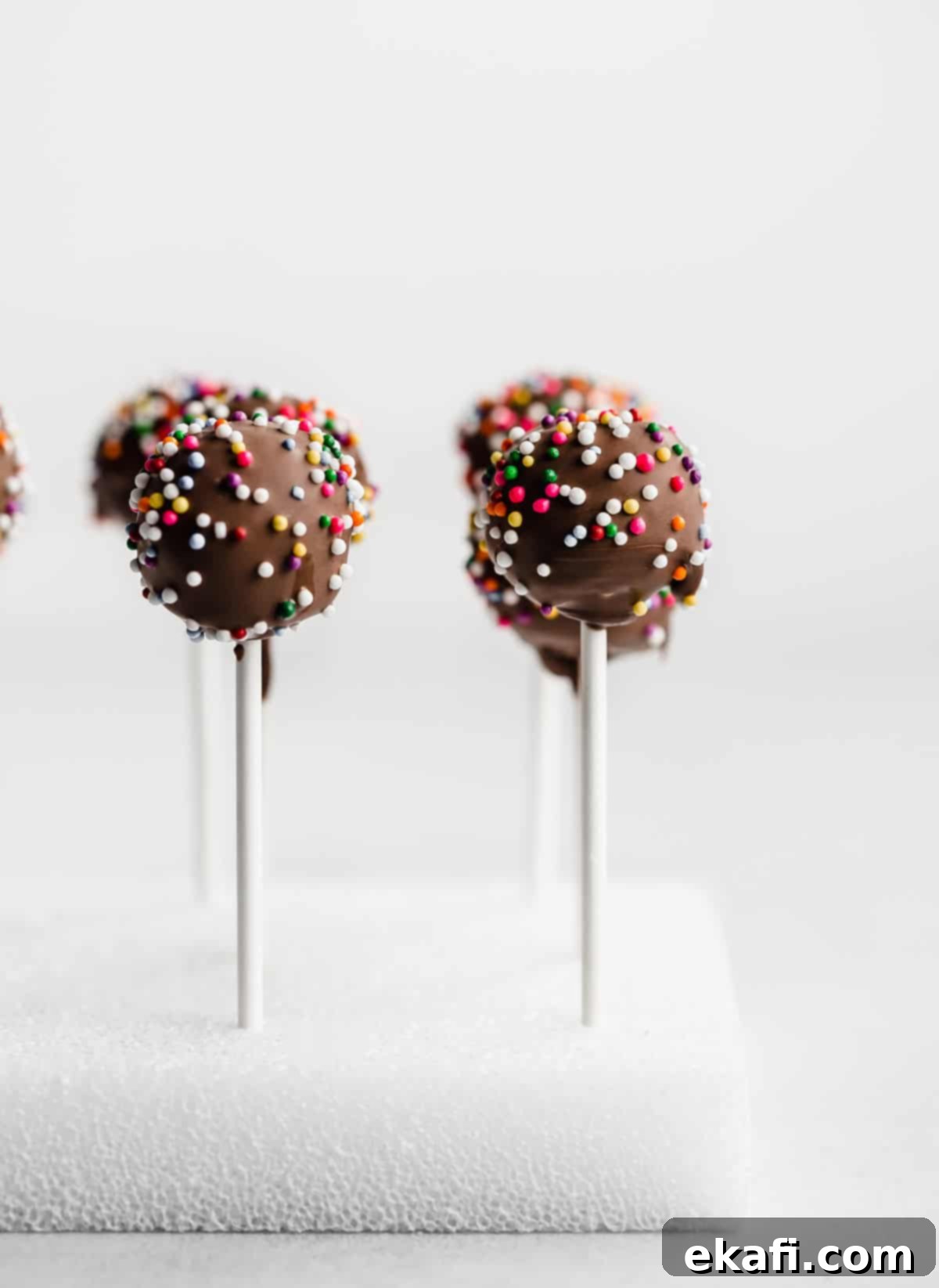 Κεκάκια Ποπ Σοκολάτας 2 Ένα σοκολατένιο cake pop με πολύχρωμη τρούφα, τοποθετημένο σε ένα λευκό κύβο από φελιζόλ σε λευκό φόντο.