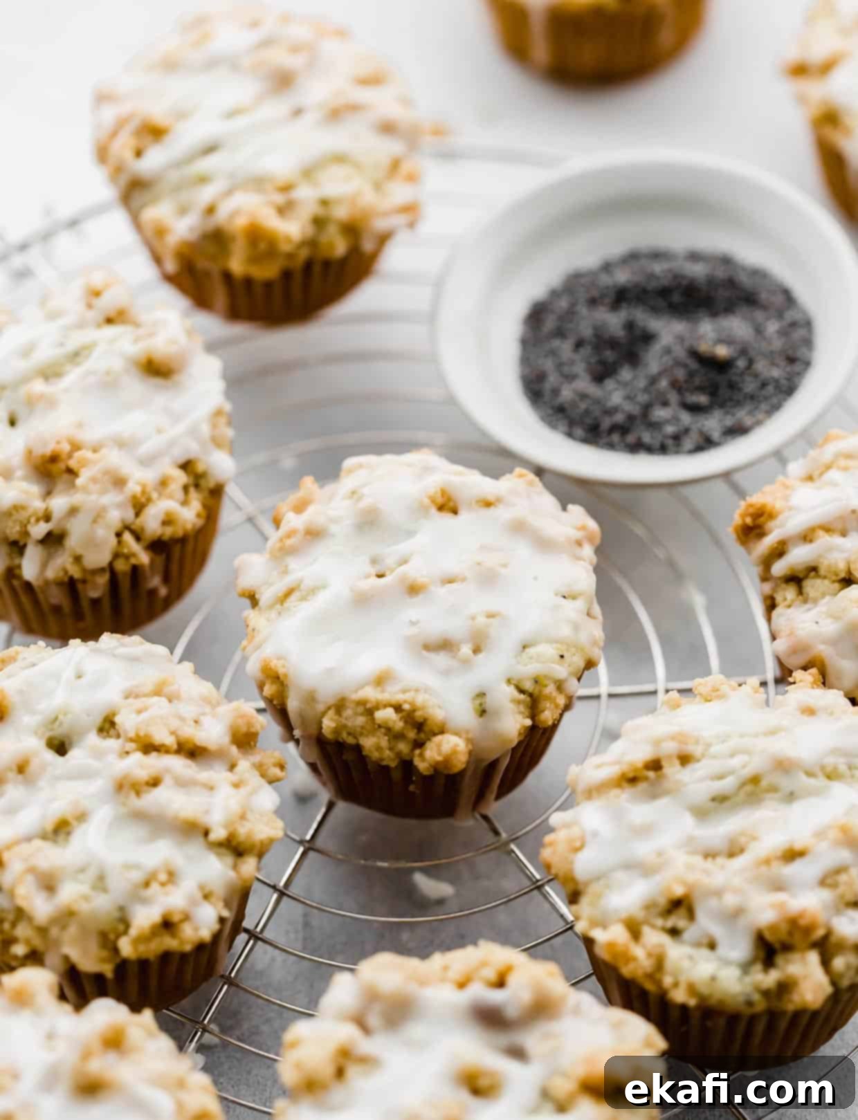Ultra Moist Lemon Poppy Seed Muffins with Sour Cream 9 A stunning arrangement of glazed and crumb-topped Lemon Poppy Seed Muffins on a wire cooling rack, with a small bowl of poppy seeds in the soft-focus background, inviting you to indulge.