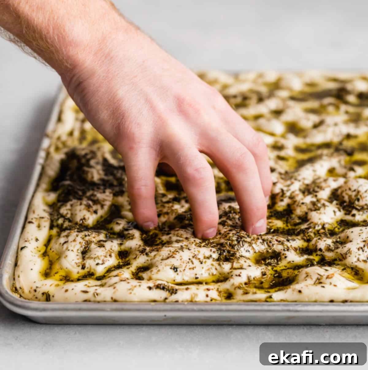 Artisanal Focaccia Bread 6 A hand pressing five fingers down into focaccia bread dough that's been topped with herb oil.