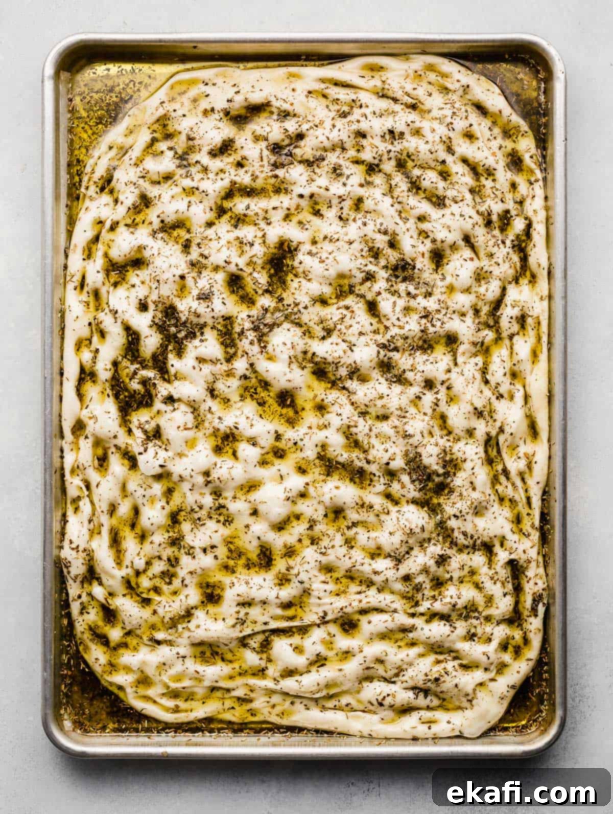 Artisanal Focaccia Bread 7 Raw focaccia bread dough dimpled and topped with herb oil on a baking sheet.