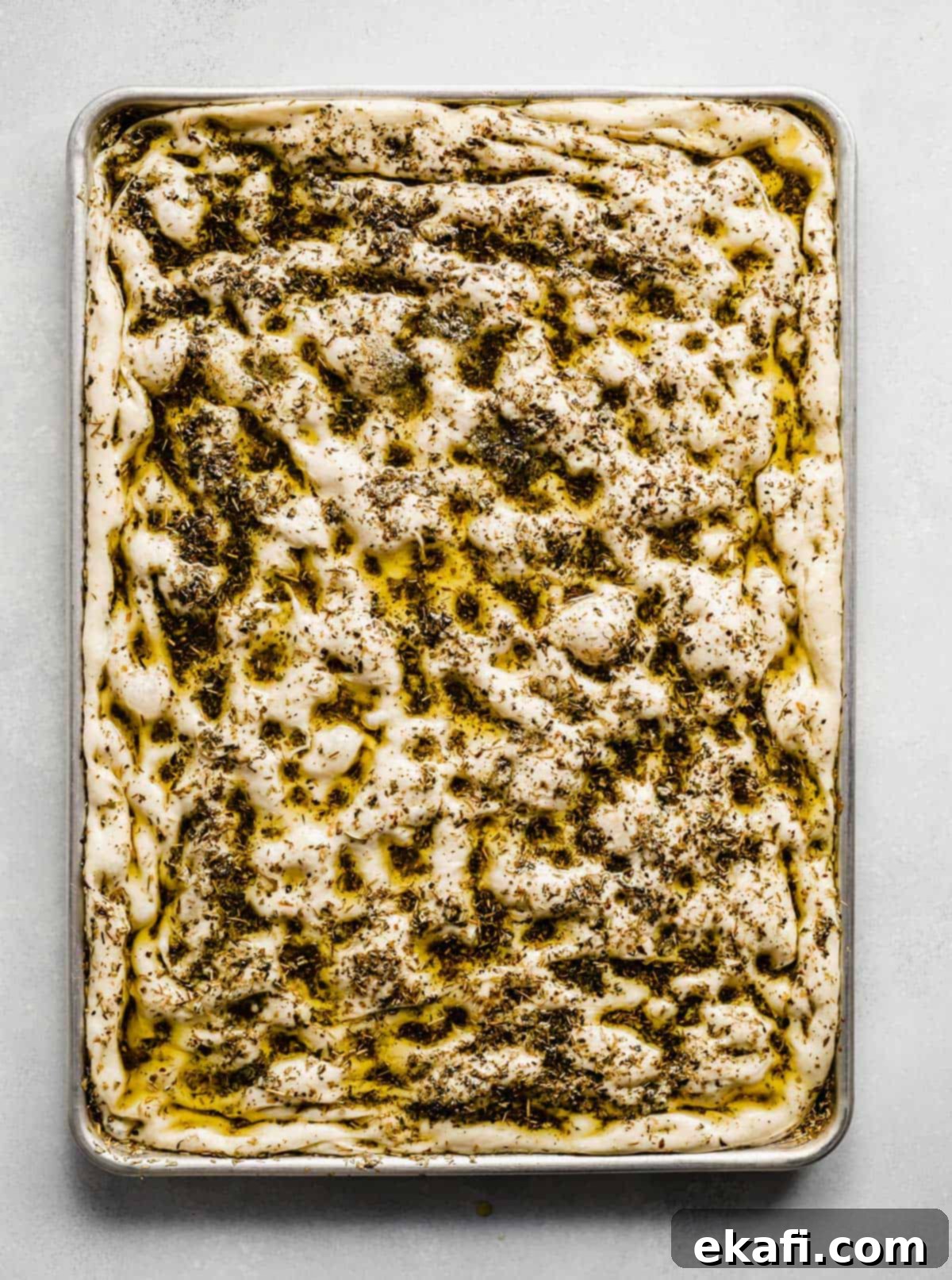 Artisanal Focaccia Bread 8 A baking sheet that has focaccia bread dough spread along the full size of the sheet, topped with herb oil.