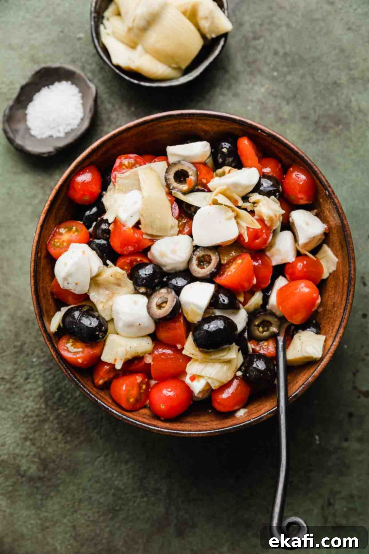 Fresh Tomato Artichoke Salad 2 A brown bowl filled with vibrant Tomato Artichoke Salad, showcasing olives and glistening mozzarella cheese balls, all set against a rich forest green background.