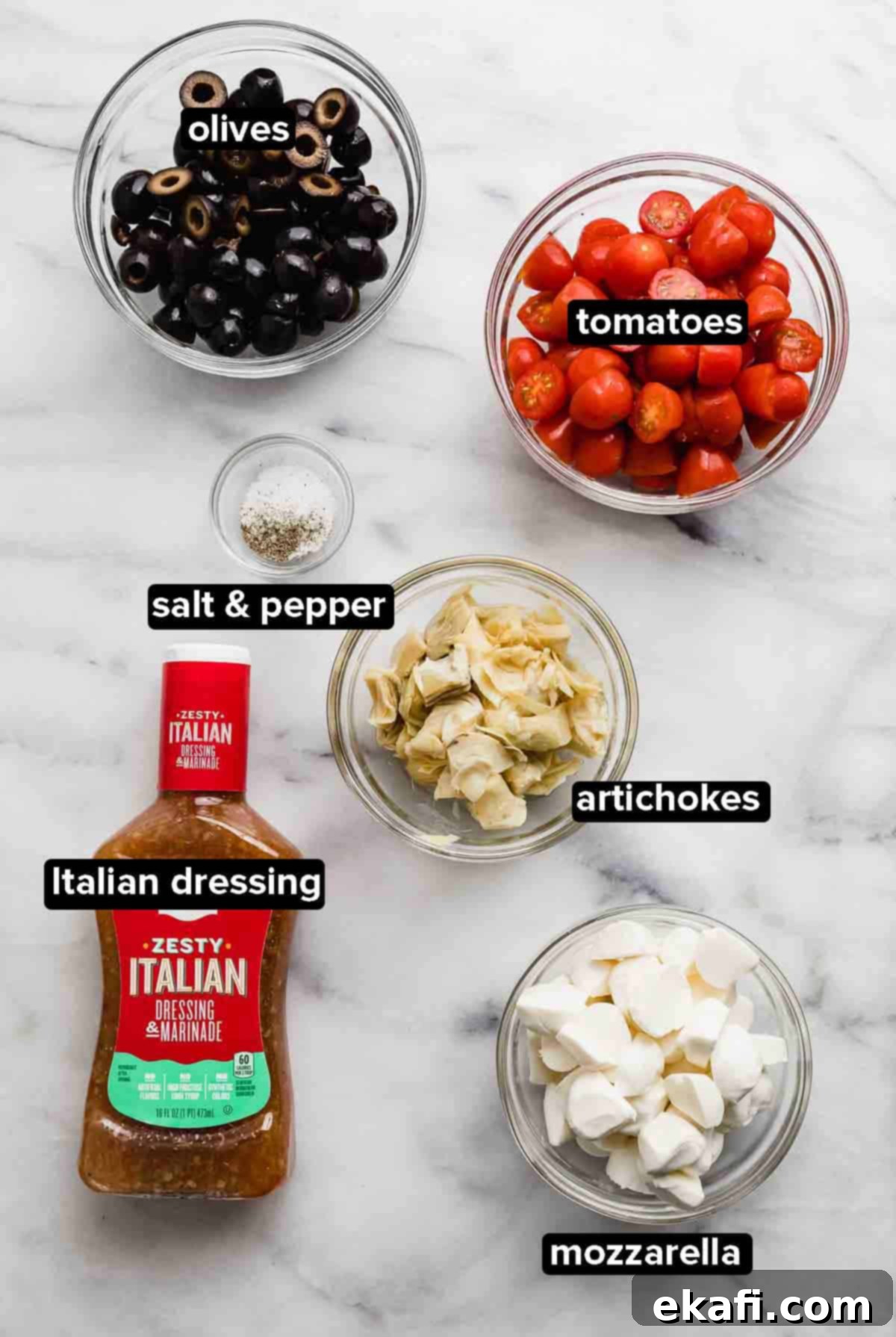 Fresh Tomato Artichoke Salad 3 A beautiful flat lay of Tomato Artichoke Salad ingredients on a clean white background, featuring black olives, a bottle of Italian dressing, fresh mozzarella pearls, vibrant tomatoes, tender artichoke hearts, and seasoning with salt and pepper.