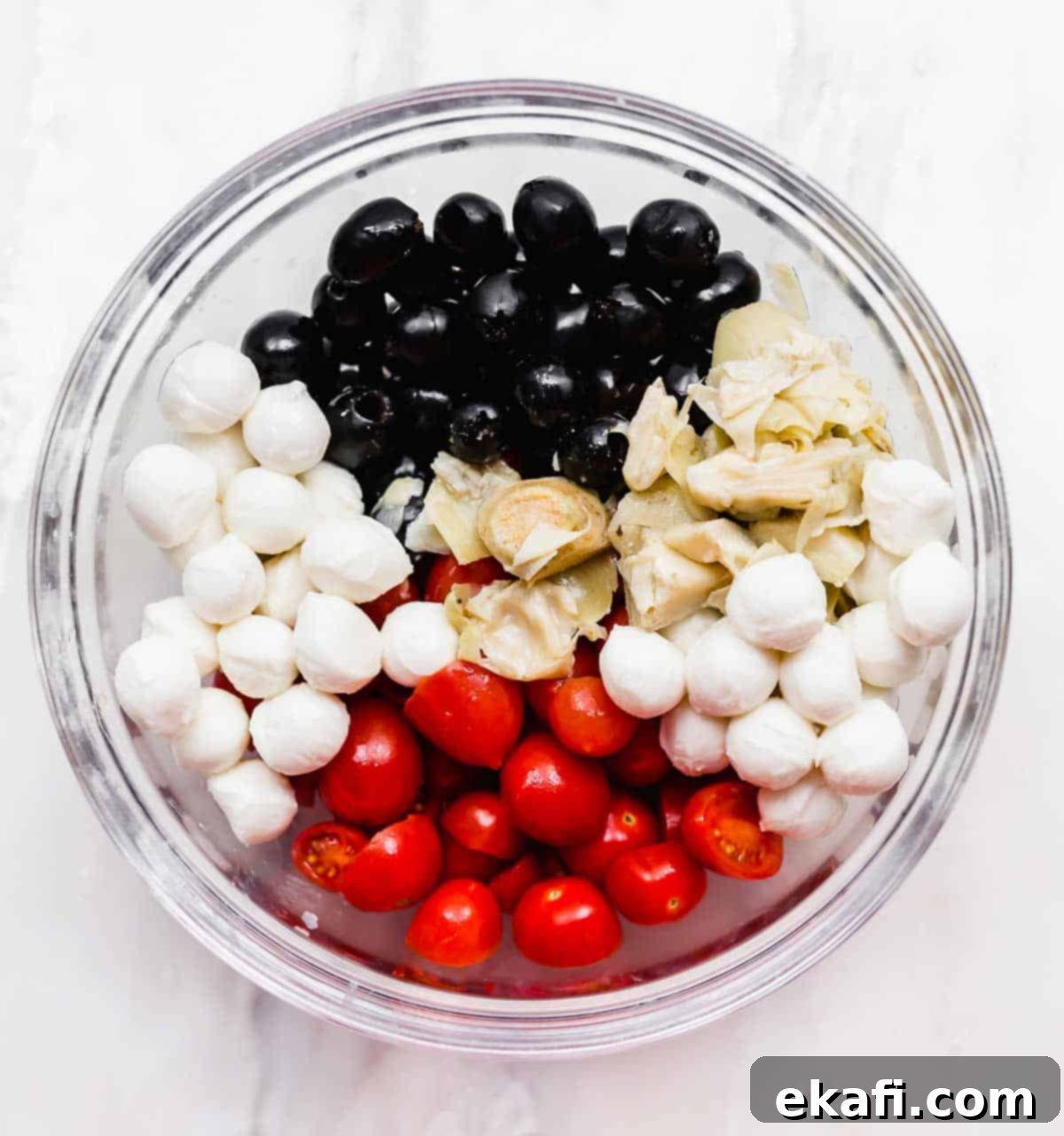 Fresh Tomato Artichoke Salad 4 A sparkling clear glass bowl filled with a delightful medley of mozzarella pearls, vibrant tomatoes, dark olives, and tender artichokes, ready to be served.