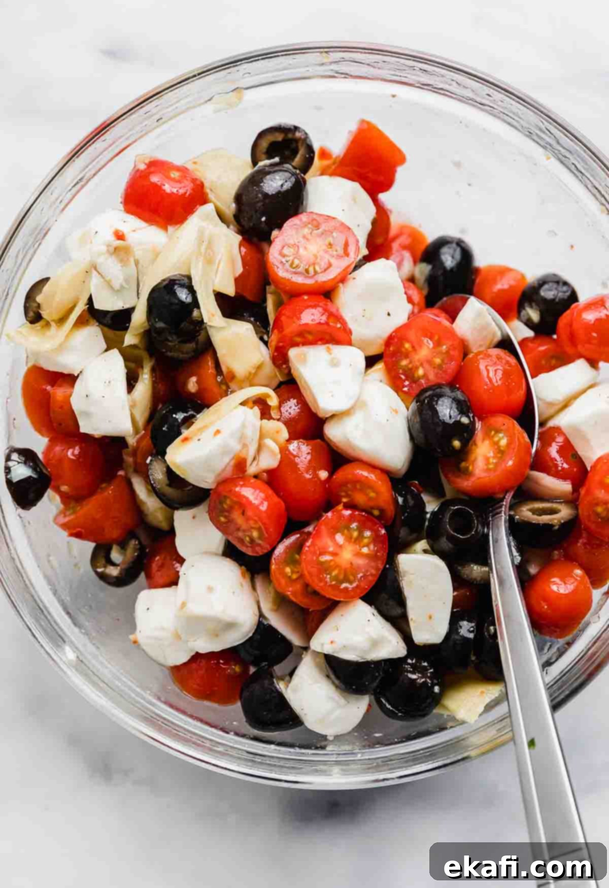 Fresh Tomato Artichoke Salad 5 A elegant glass bowl showcases a beautifully arranged Mozzarella, Olive, and Tomato Artichoke Salad, sitting on a pristine white background.