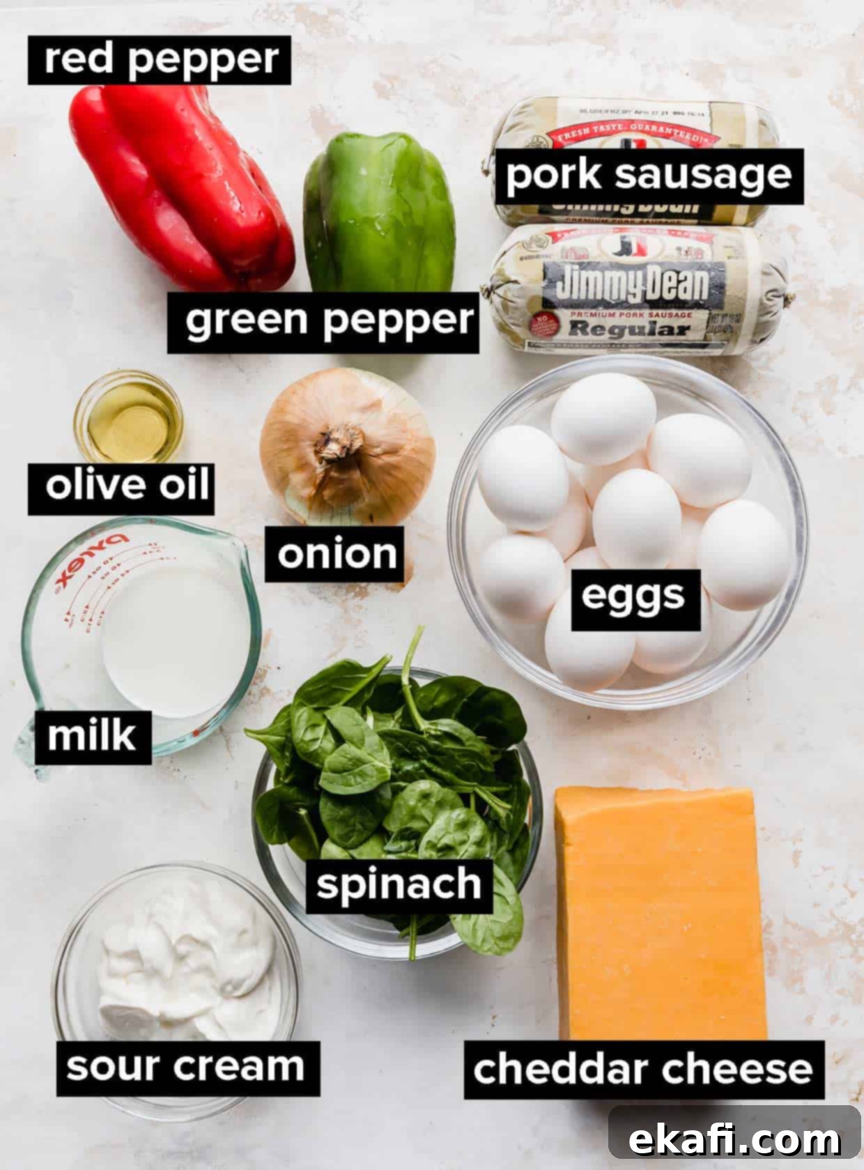 Effortless Breakfast Casserole 3 A selection of fresh ingredients laid out on a textured white background, including eggs, sausage, spinach, peppers, onion, sour cream, milk, and cheese.