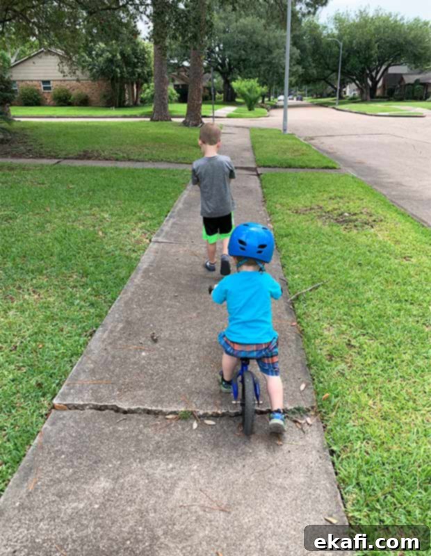 Two young boys on a sidewalk, one on a strider bike and the other walking beside him, enjoying their morning walk.
