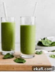 Πράσινο Πρωτεϊνούχο Smoothie 8 Δύο ποτήρια γεμάτα με ένα πράσινο smoothie πρωτεΐνης σε ξύλινη επιφάνεια κοπής με ένα μπολ σπανάκι στο βάθος.