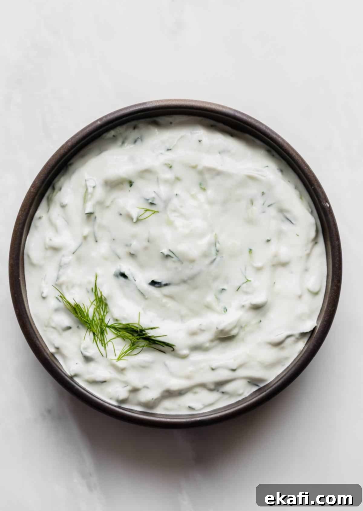 Authentic Tzatziki Recipe 2 A black rimmed bowl filled with authentic Tzatziki sauce using shredded cucumber and fresh dill, on a white background.