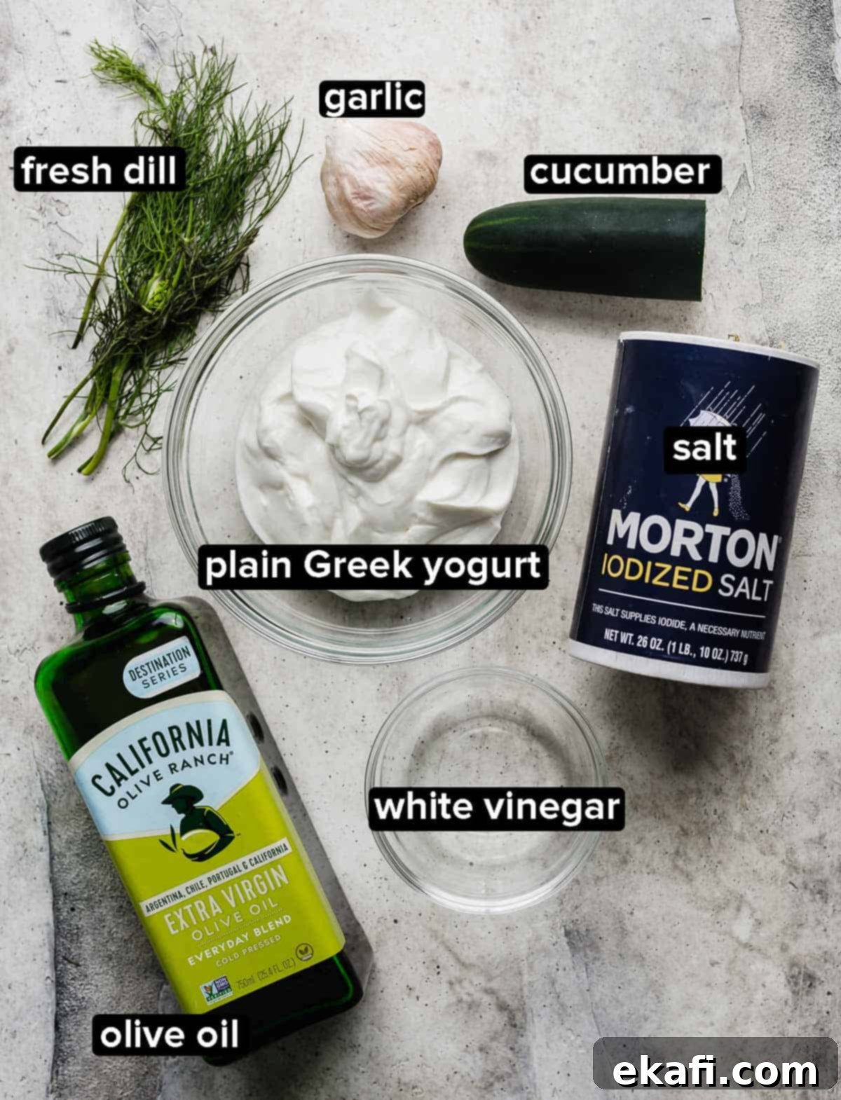 Authentic Tzatziki Recipe 3 Tzatziki ingredients on a light gray background: salt, cucumber, dill, olive oil, greek yogurt, garlic, and vinegar.