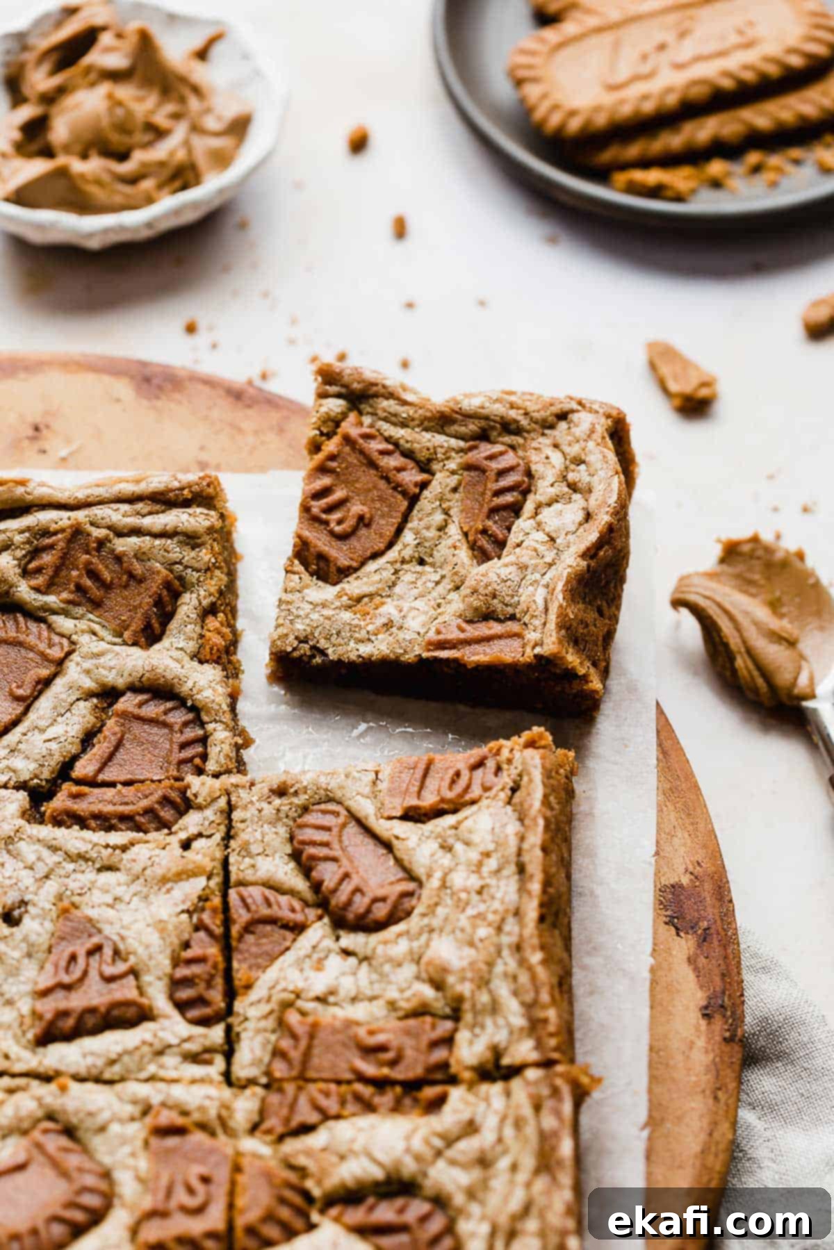 Μπράουνις Λωτού 7 A square of Biscoff Brownies on a white parchment paper with Biscoff cookies in the background.