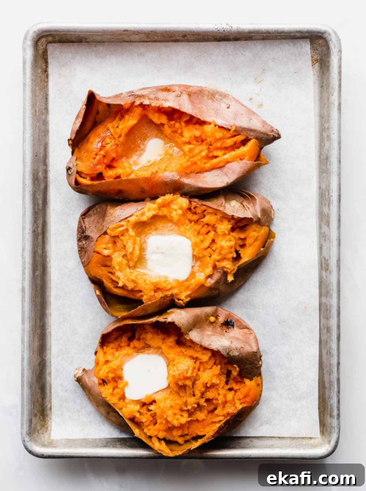 Easy Baked Sweet Potato 6 Three baked sweet potatoes on a baking sheet, each cut open with a square of butter in the orange flesh.